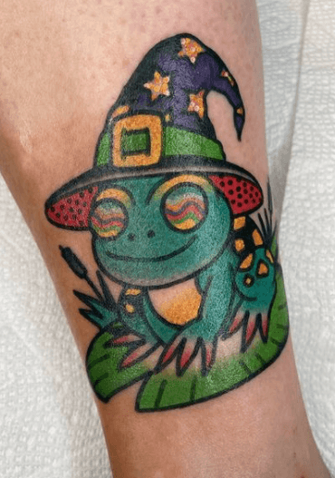 Tattoo of a smiling frog on a lily pad wearing a wizard hat.