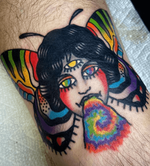 A tattoo of a woman's head with moth wings shooting a rainbow from her mouth.